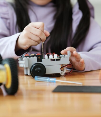 A young girl concentrated on creating a robot surrounded by tools and electronic components.